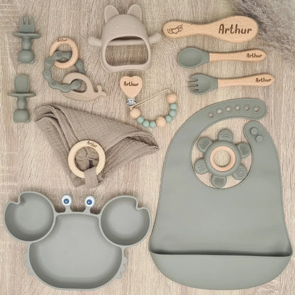 Vue détaillée des 10 pièces du coffret naissance personnalisé : bavoir, couverts en bois gravés, serviette et jouets de dentition en silicone.
