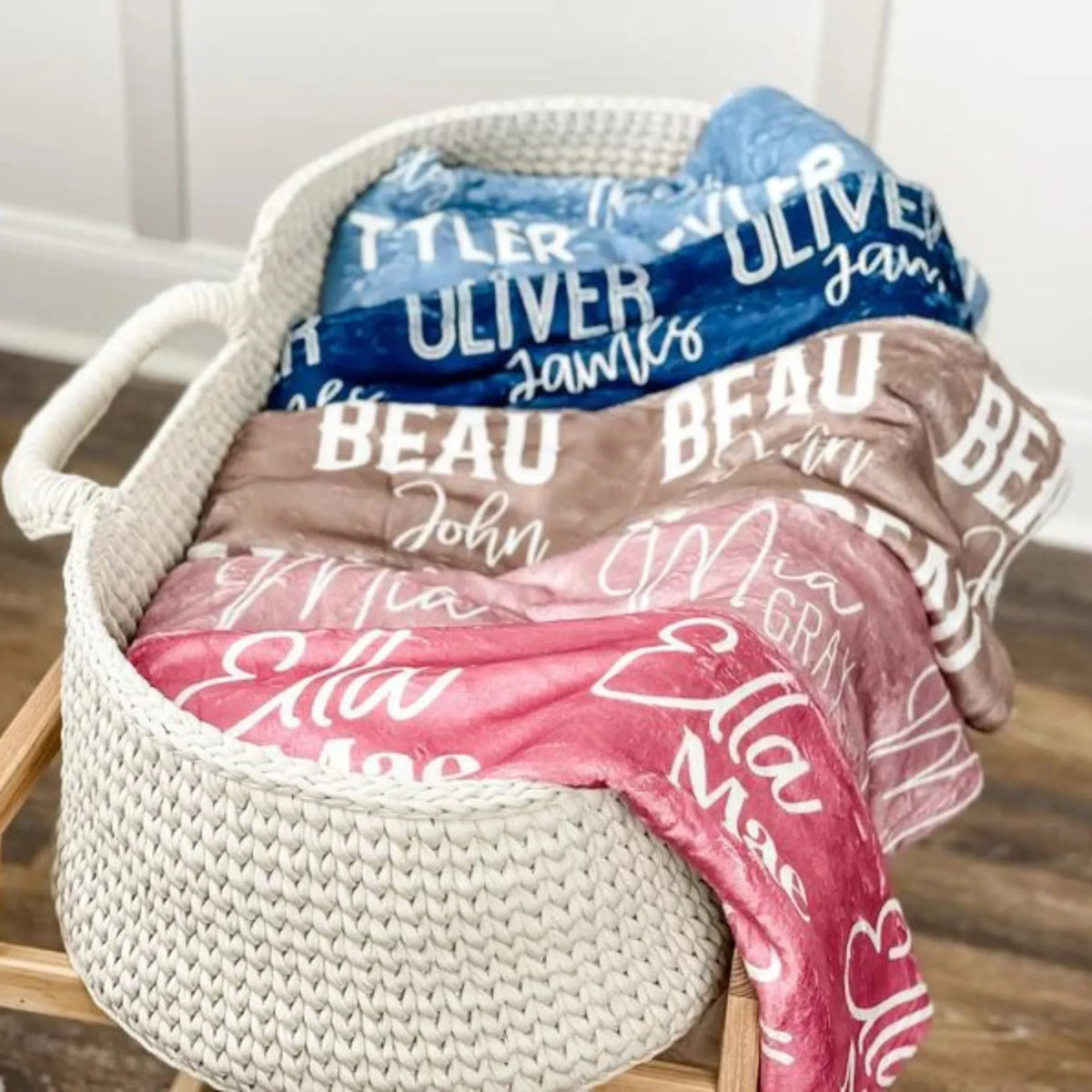 Panier tressé rempli de couvertures pliées, mettant en avant un plaid personnalisé bleu, beige et rose avec différentes typographies.