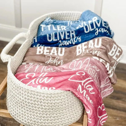 Panier tressé rempli de couvertures pliées, mettant en avant un plaid personnalisé bleu, beige et rose avec différentes typographies.