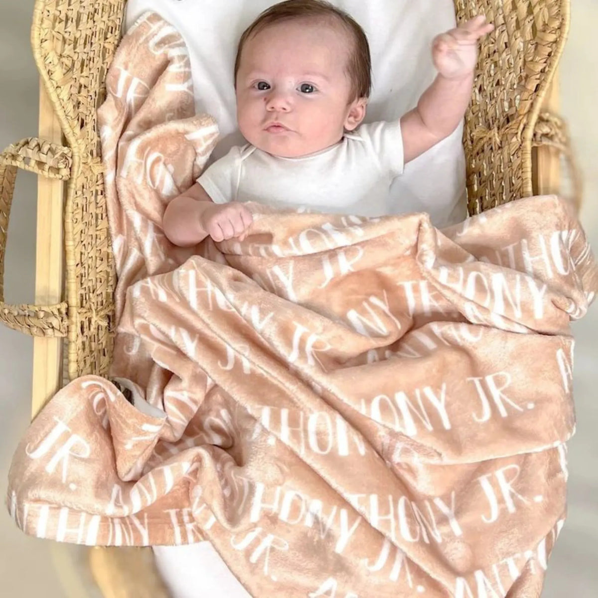 Bébé éveillé couché dans un couffin en osier naturel, posé sur un plaid personnalisé couleur sable ou beige avec le nom Anthony Jr.