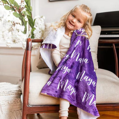 Petite fille souriante assise dans un fauteuil, s'enveloppant confortablement dans un plaid personnalisé de couleur violette au prénom Maya.