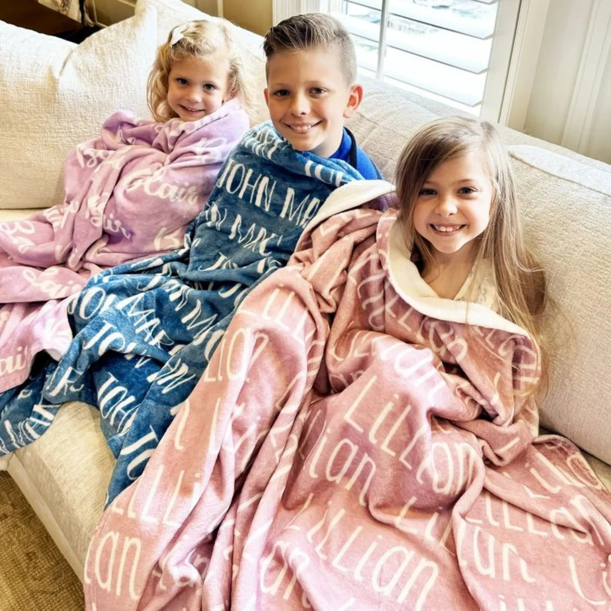 Trois enfants sur un canapé beige avec leurs couvertures : un plaid personnalisé lilas, un bleu et un vieux rose.