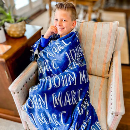 Garçon souriant assis dans un fauteuil rayé, tenant un plaid personnalisé bleu foncé avec typographie blanche.