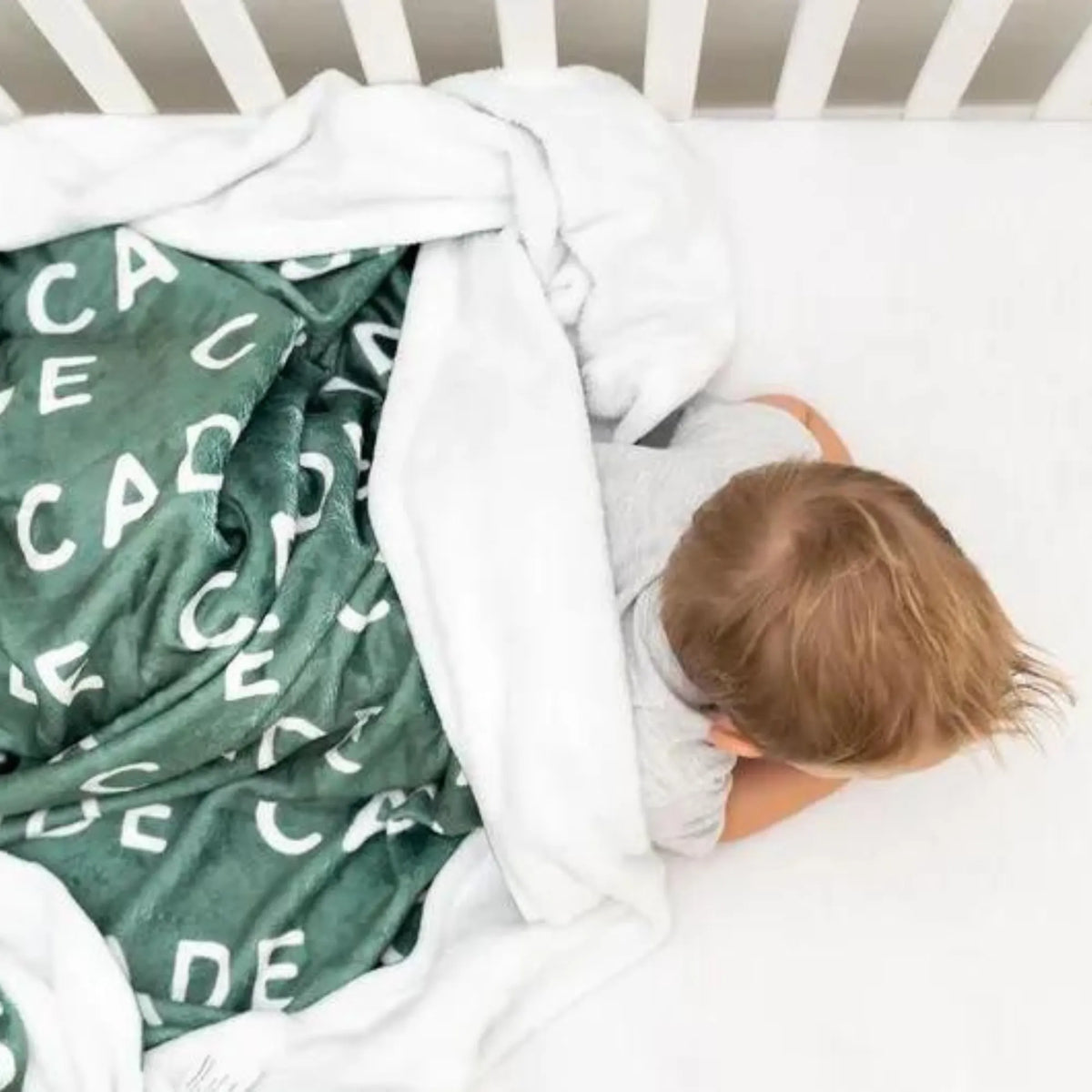 Jeune enfant faisant la sieste dans un lit à barreaux blancs, bordé par un plaid personnalisé vert sapin avec le prénom Cade.