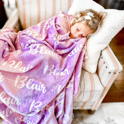 Petite fille dormant profondément dans un fauteuil, blottie sous un plaid personnalisé couleur lilas ou parme.