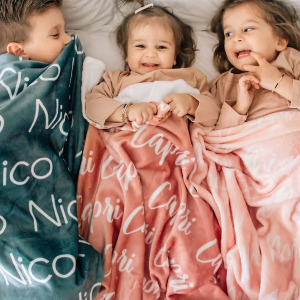Trois enfants allongés têtes contre têtes, couverts par un plaid personnalisé vert canard, terracotta et rose pâle.