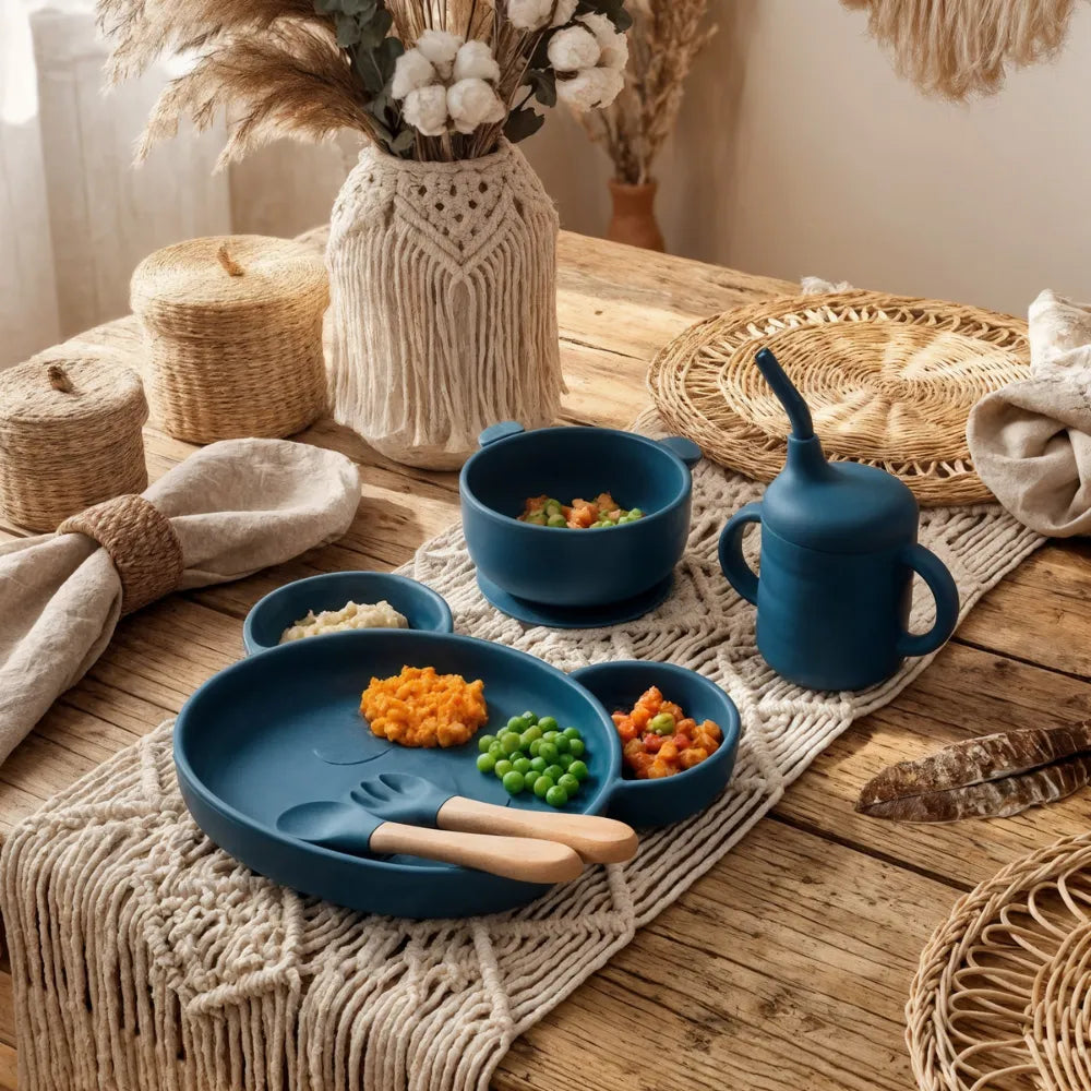 Set complet vaisselle bébé en silicone bleu marine Baby-Crea. Comprend assiette compartimentée ventouse, bol, tasse d'apprentissage et couverts, servis avec repas pour DME sur une table bohème.